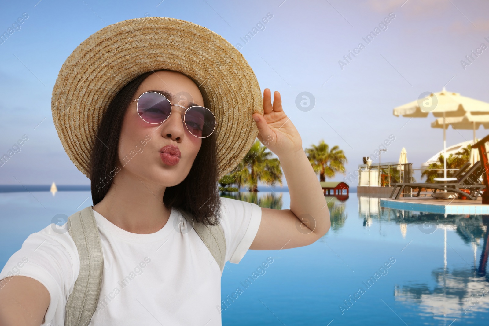 Image of Smiling young woman in sunglasses and straw hat taking selfie at resort