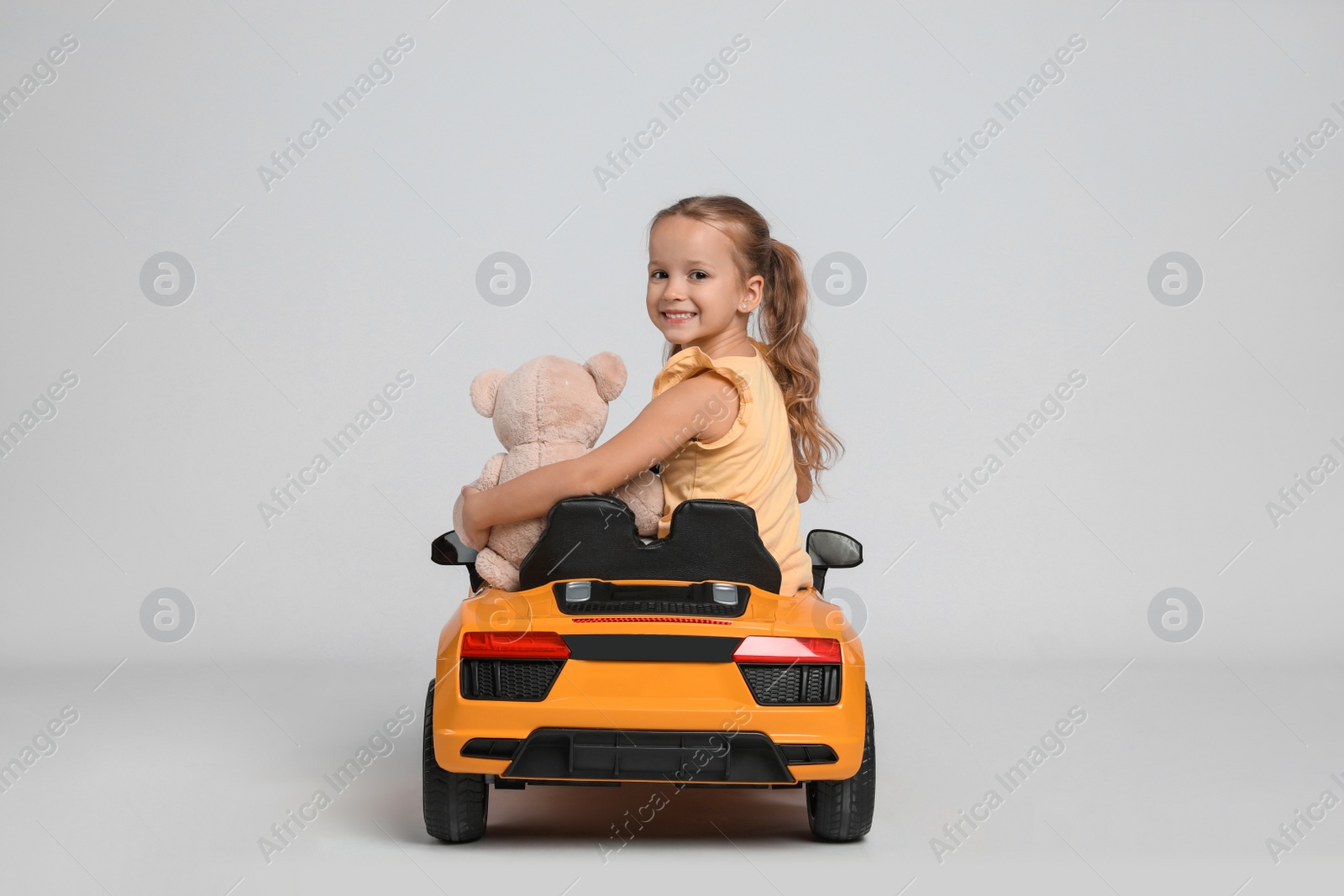 Photo of Cute little girl with toy bear driving children's car on grey background