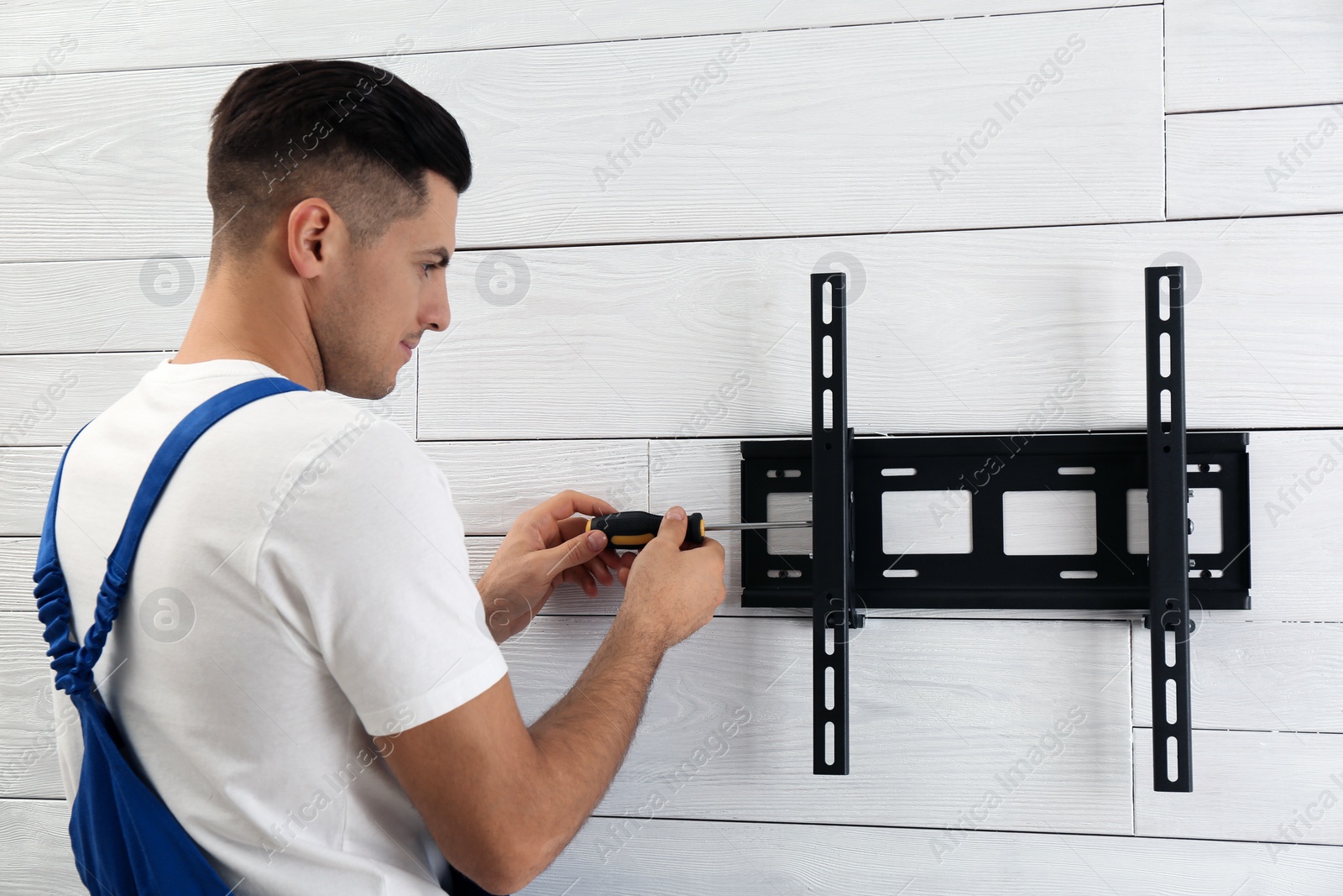 Photo of Professional technician with screwdriver installing TV bracket on wall indoors