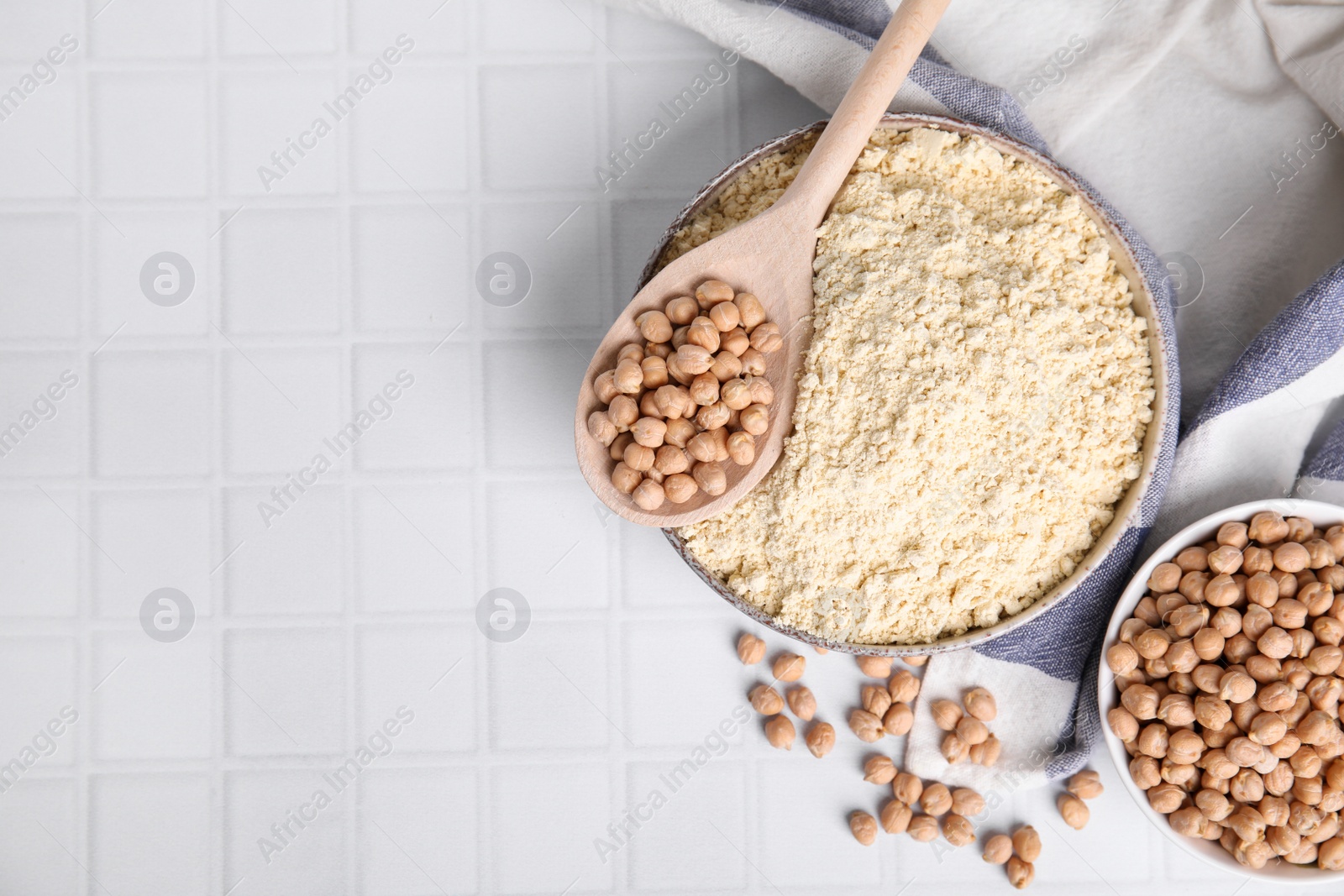 Photo of Chickpea flour in bowl and seeds on white tiled table, flat lay. Space for text