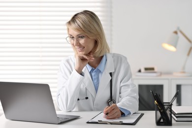 Smiling doctor with laptop having online consultation at table in office