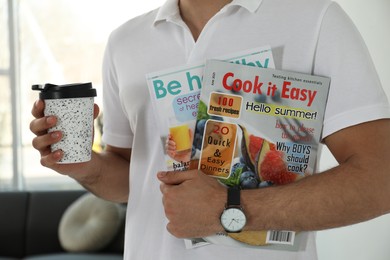 Photo of Man holding cup of coffee and different magazines at home, closeup