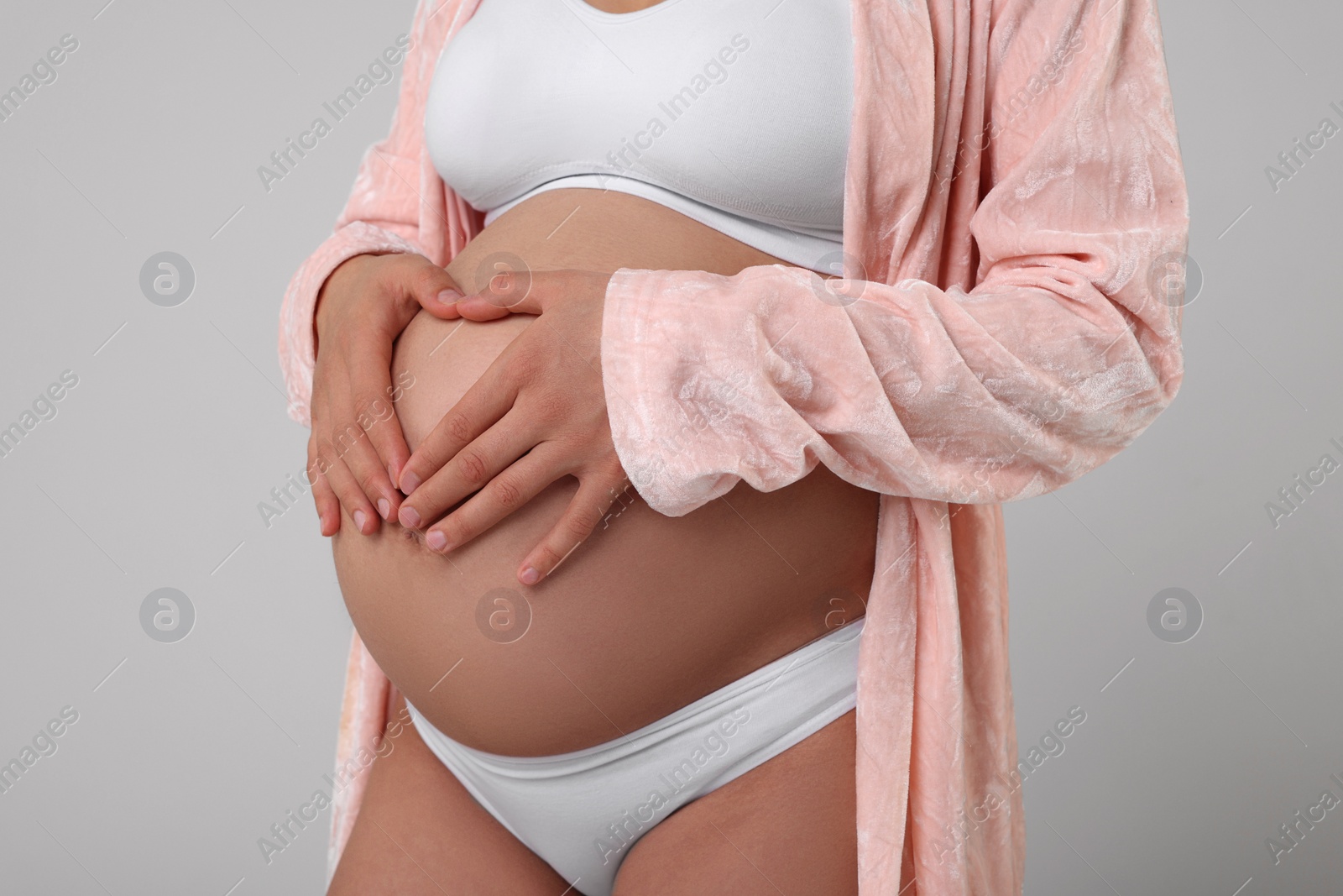 Photo of Pregnant woman in stylish comfortable underwear and robe making heart with hands on her belly against grey background, closeup