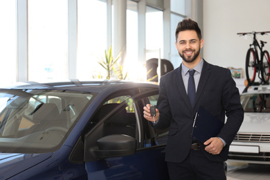 Photo of Young salesman with key and clipboard near car in dealership