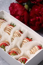 Photo of Delicious chocolate covered strawberries in box and flowers on light grey table, closeup