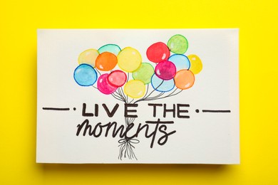 Photo of Card with life-affirming phrase Live The Moments on yellow background, top view