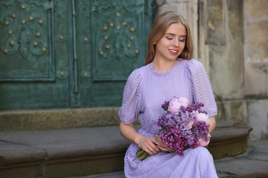 Beautiful woman with bouquet of spring flowers near building outdoors, space for text