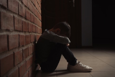 Photo of Sad little boy sitting on floor near brick wall indoors