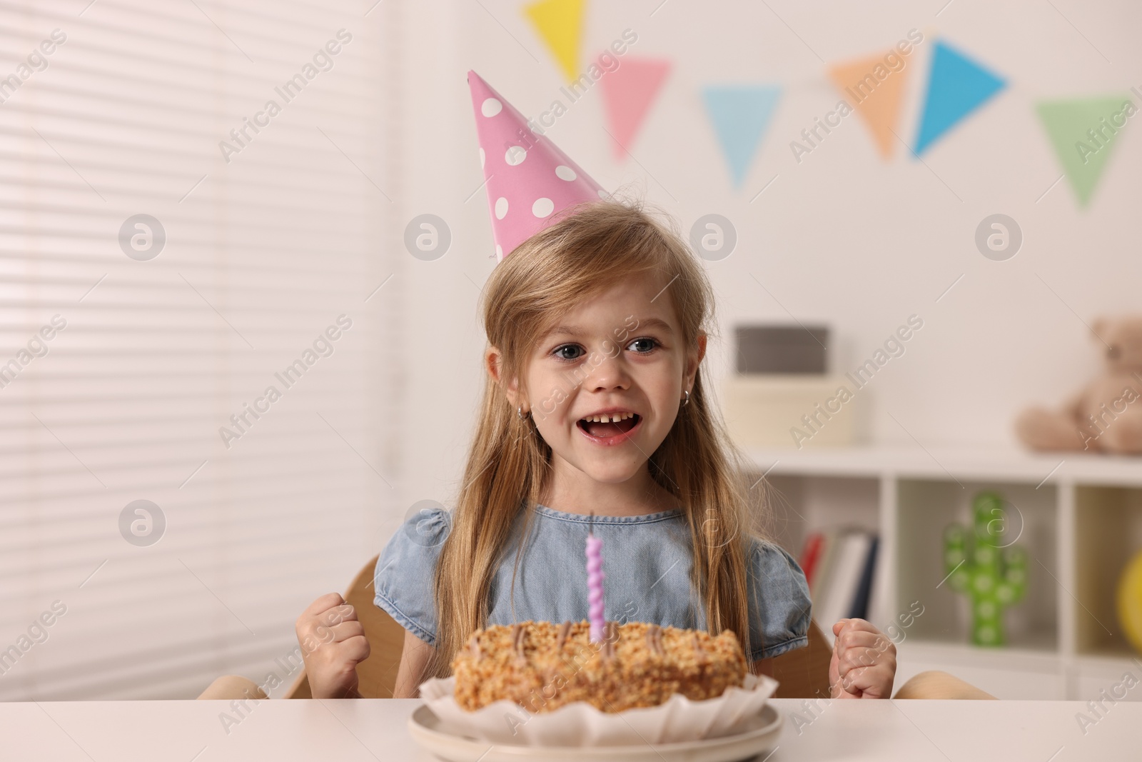 Photo of Cute girl in party hat with birthday cake at table indoors