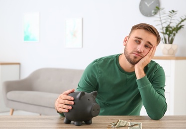 Photo of Young man with piggy bank and money at table indoors. Space for text