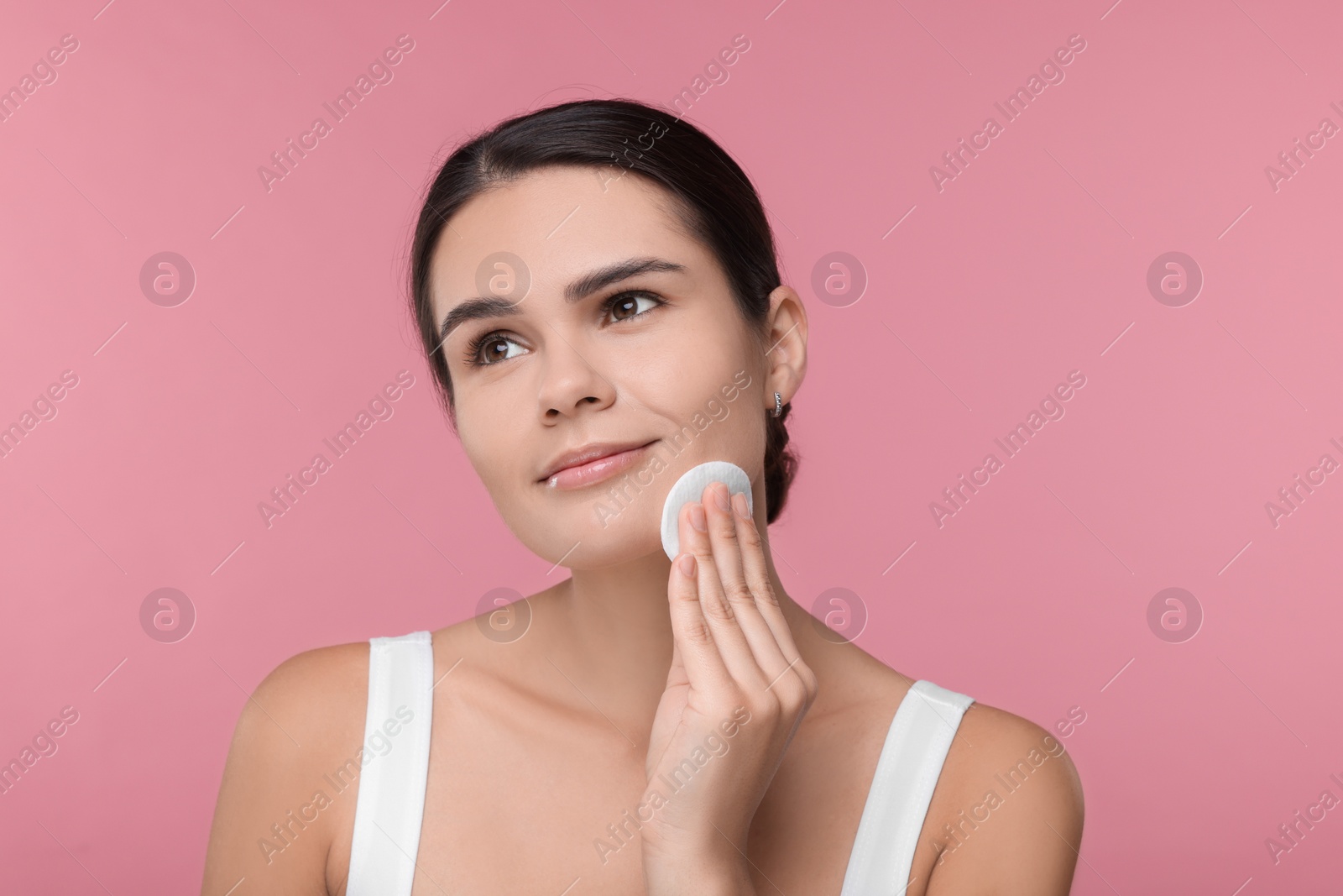 Photo of Young woman cleaning her face with cotton pad on pink background