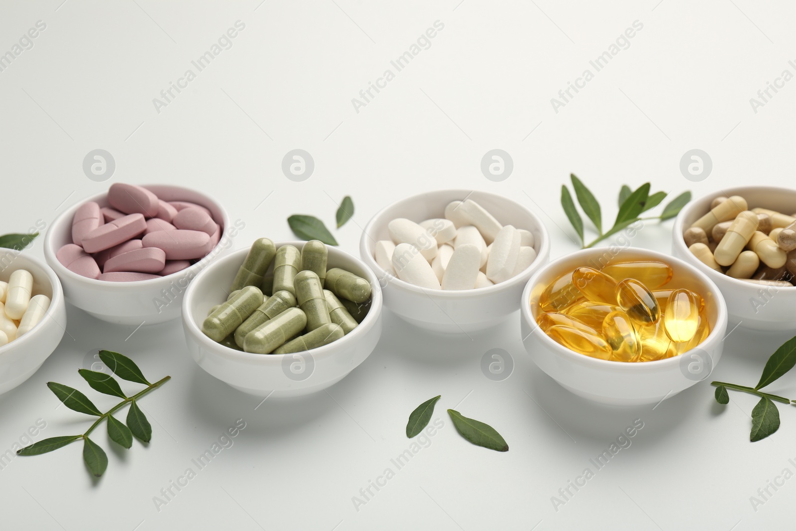 Photo of Different vitamin capsules in bowls and leaves on white background, closeup