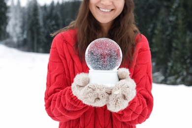 Photo of Woman with knitted mittens holding snow globe outdoors, closeup. Winter vacation