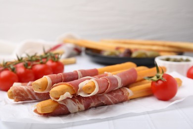 Photo of Delicious grissini sticks with prosciutto and snacks on white table, closeup