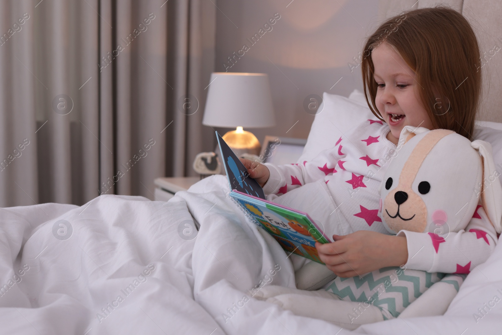 Photo of Cute little girl with toy reading book in bed at home