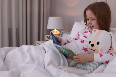 Cute little girl with toy reading book in bed at home