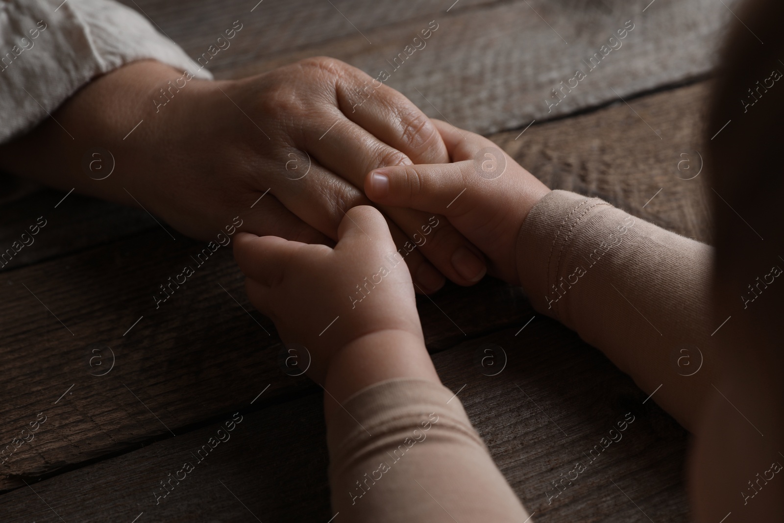 Photo of Woman holding hands with her granddaughter at wooden table, closeup