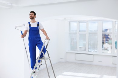 Photo of Handyman with roller on step ladder in room. Ceiling painting