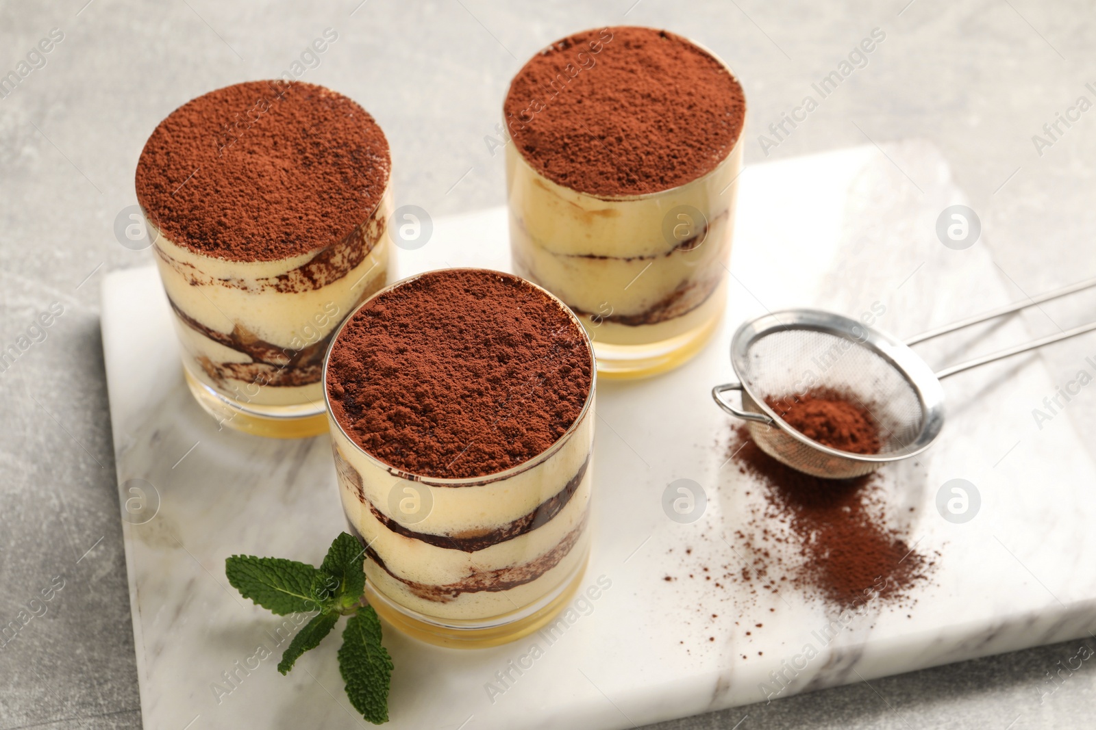 Photo of Delicious tiramisu in glasses, mint and sieve with cocoa powder on grey textured table, above view