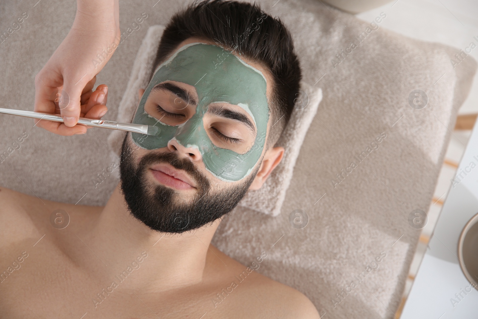 Photo of Cosmetologist applying mask on man's face in spa salon, top view