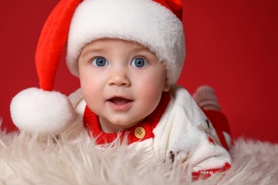 Photo of Cute baby in Santa hat on fluffy carpet against red background, closeup. Christmas celebration