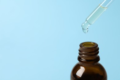 Photo of Dripping cosmetic oil from pipette into bottle on light blue background, closeup. Space for text