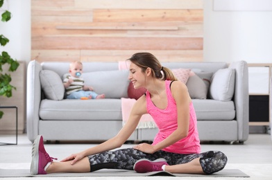 Photo of Young sportive woman doing exercise while her son sitting on sofa at home. Fitness training