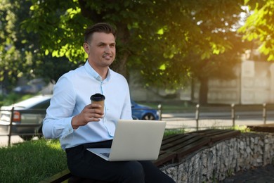 Photo of Handsome man with cup of coffee using laptop on bench outdoors. Space for text