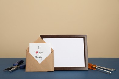 Photo of Happy Father's Day. Card with phrase I Love You, Daddy in envelope, tools and empty frame on blue table. Space for text