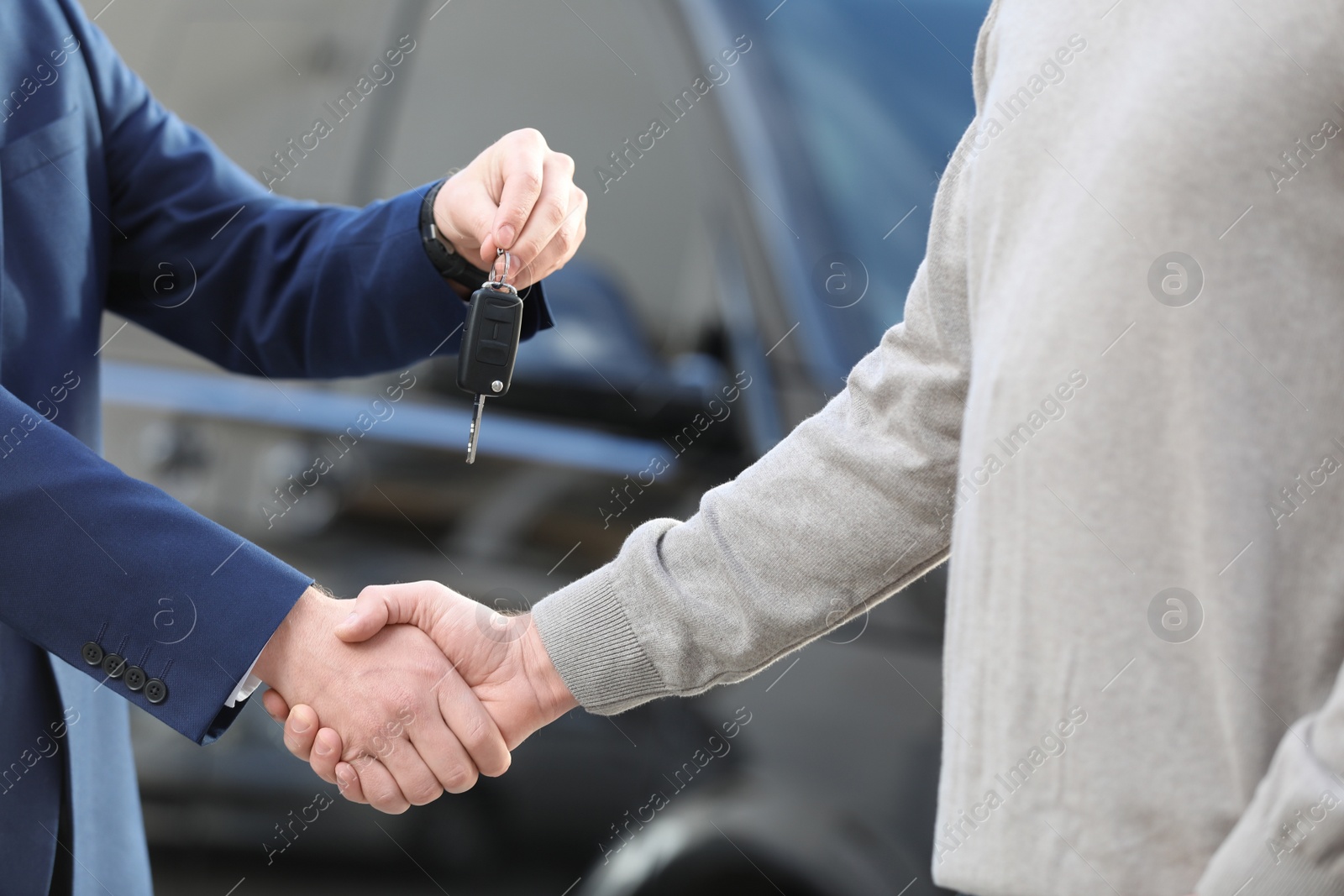 Photo of Salesman giving key to customer while shaking hands in modern auto dealership, closeup. Buying new car