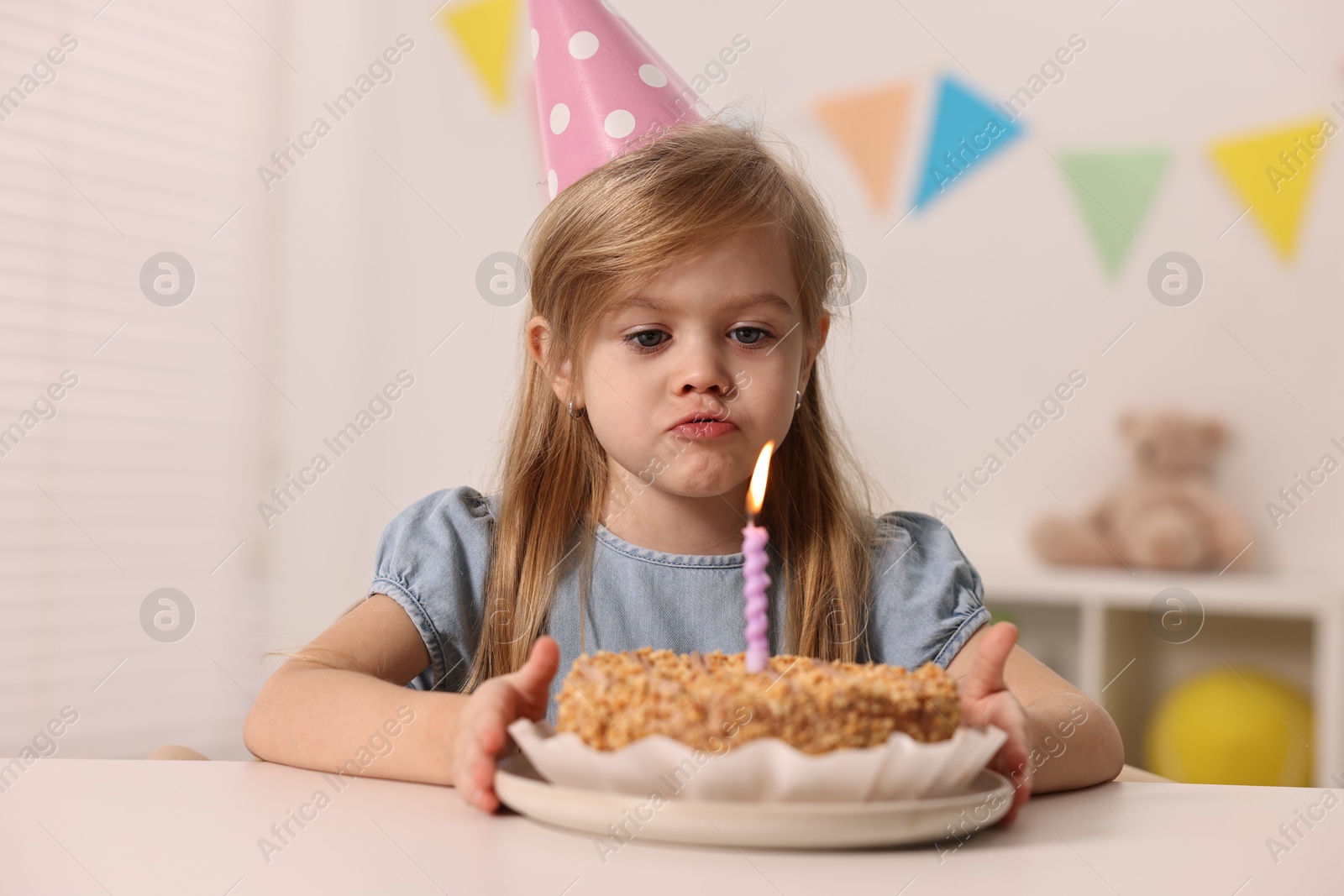 Photo of Cute girl in party hat with birthday cake at table indoors