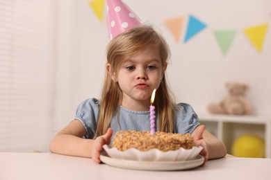 Cute girl in party hat with birthday cake at table indoors