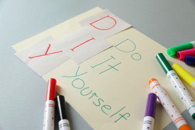 Photo of Sheet with written phrase Do It Yourself and colorful markers on grey background. DIY concept