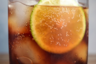 Glass of refreshing soda drink with ice cubes and lime on blue background, closeup