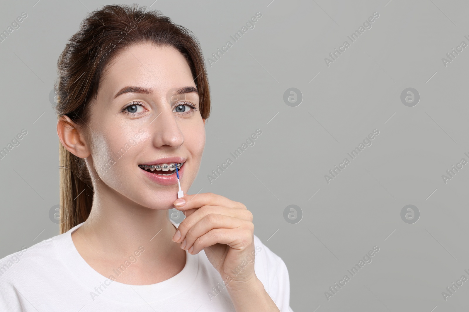 Photo of Smiling woman with dental braces cleaning teeth using interdental brush on grey background. Space for text