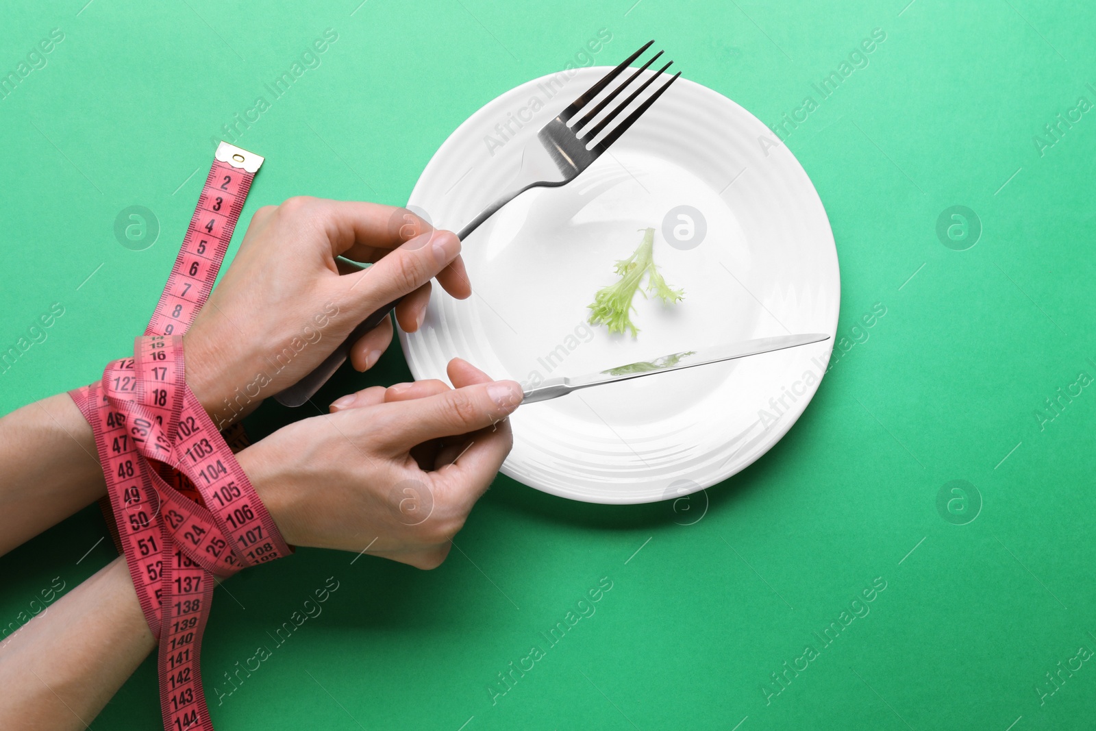 Photo of Diet concept. Woman holding cutlery in hands tied with measuring tape over plate with lettuce leaf on green background, top view