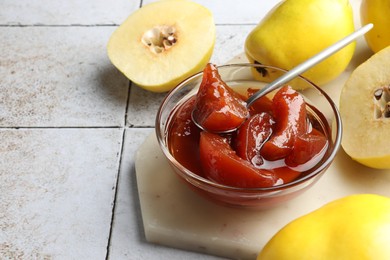 Tasty homemade quince jam in bowl, spoon and fruits on tiled table, closeup. Space for text