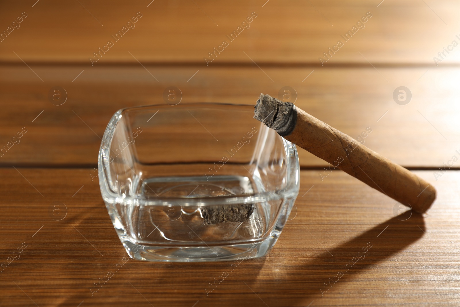 Photo of Smoldering cigar near glass ashtray on wooden table, closeup