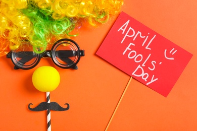 Funny face made of clown's accessories and sign with phrase April Fools' Day on red background, flat lay