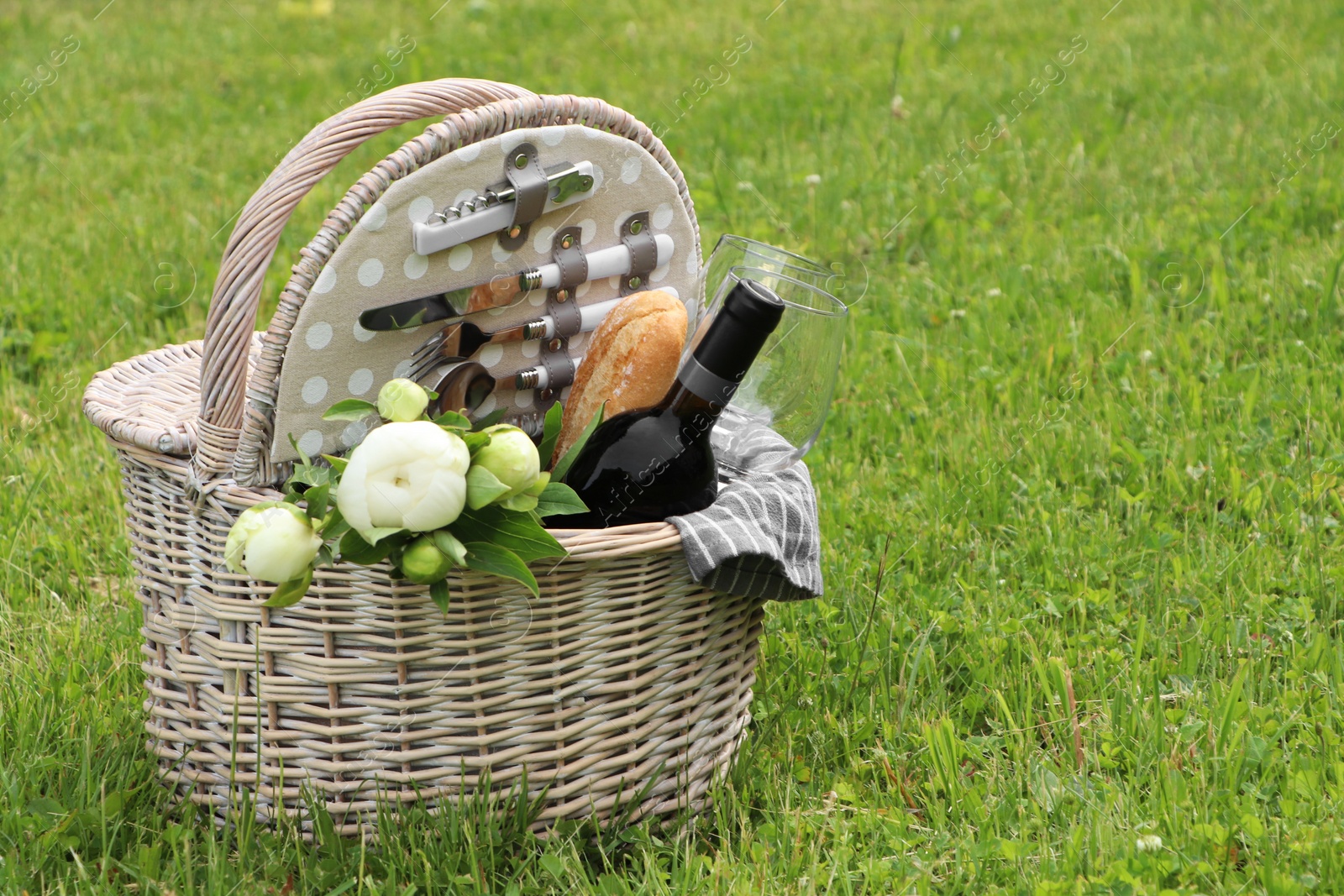 Photo of Picnic basket with wine, bread and flowers on green grass outdoors