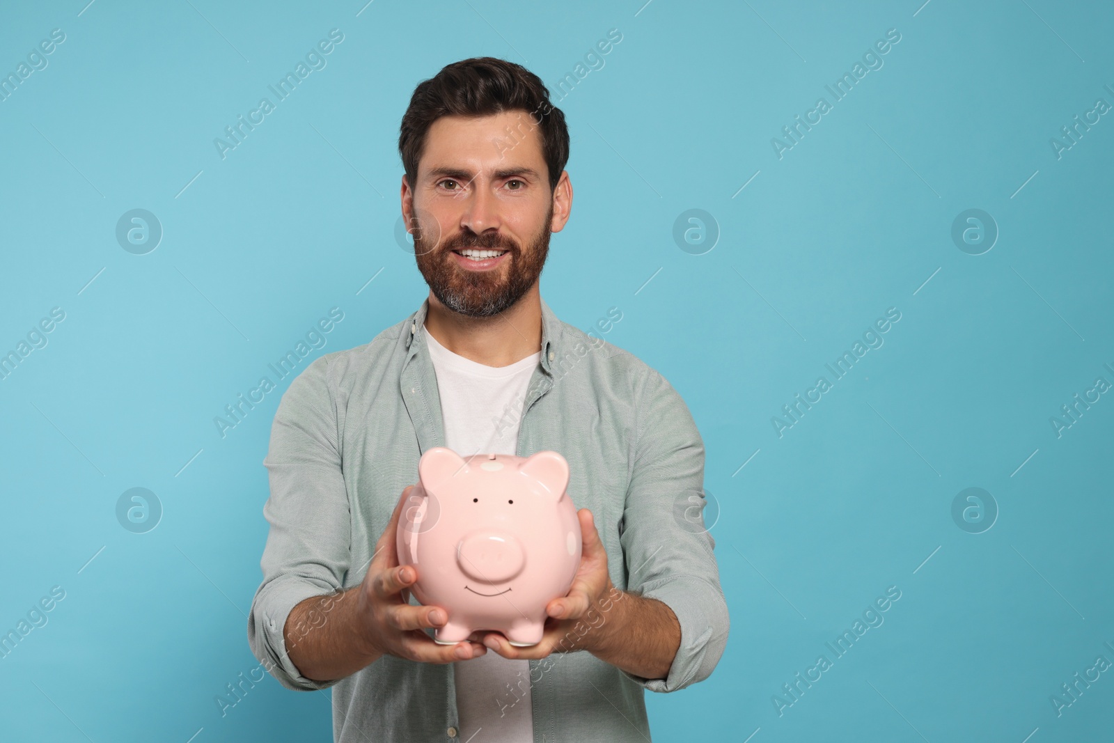 Photo of Happy man with ceramic piggy bank on light blue background, space for text