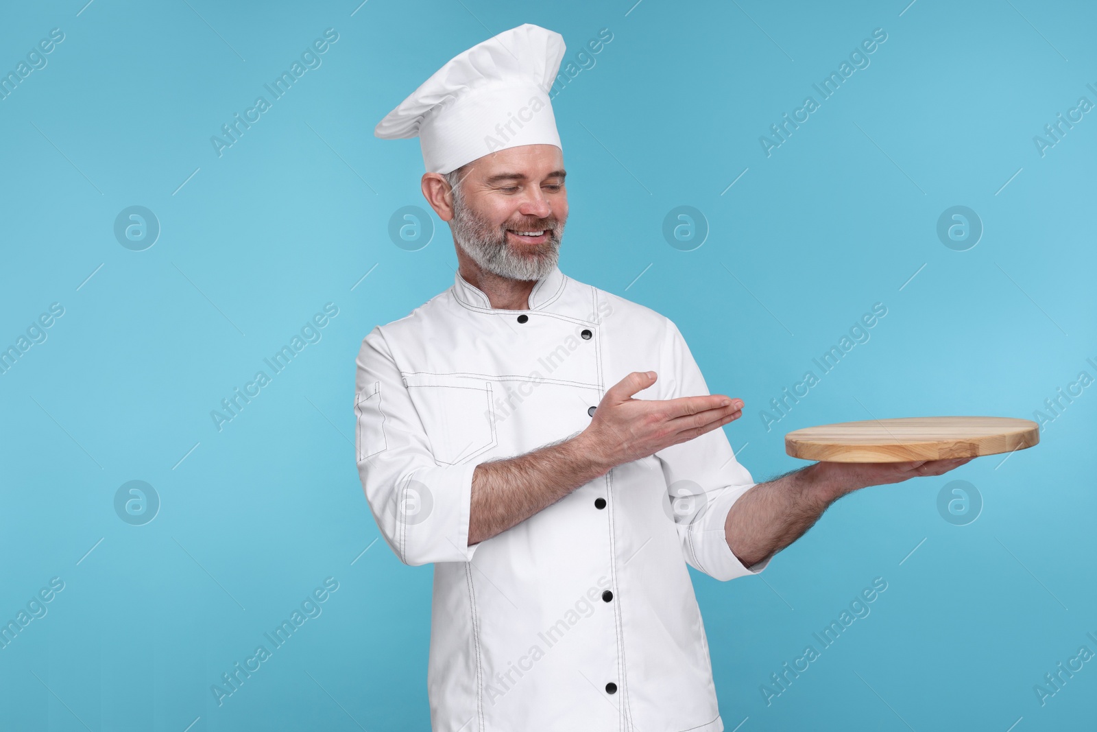 Photo of Happy chef in uniform showing wooden board on light blue background