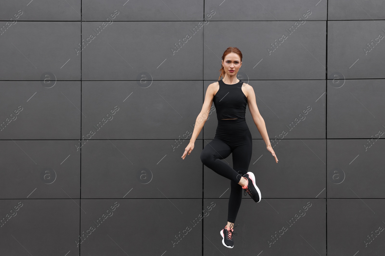 Photo of Beautiful woman in gym clothes jumping near dark grey wall on street, space for text