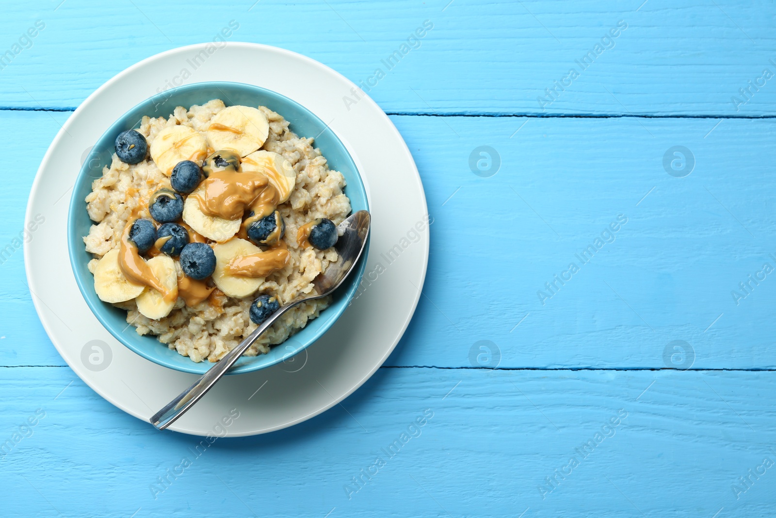 Photo of Tasty oatmeal with banana, blueberries and peanut butter served in bowl on light blue wooden table, top view. Space for text