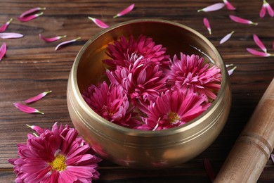 Tibetan singing bowl with water, beautiful flowers and mallet on wooden table, closeup