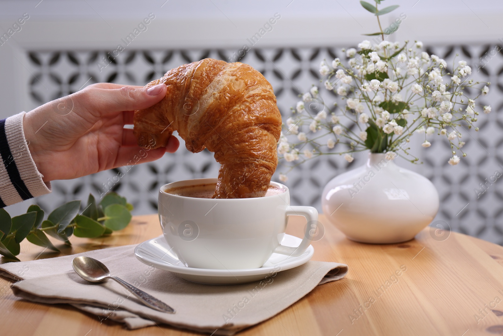 Photo of Tasty break. Woman dipping fresh croissant into cup with cappuccino at wooden table indoors, closeup