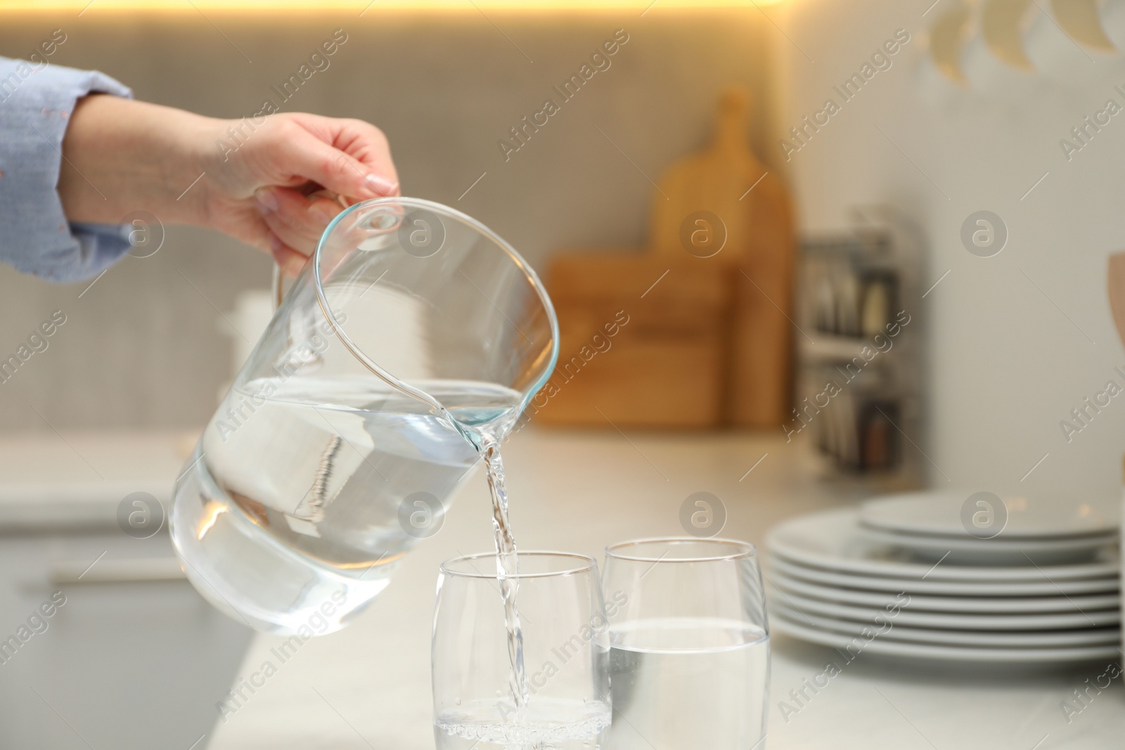 Photo of Woman pouring water from jug into glass at white table in kitchen, closeup. Space for text