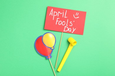Sign with phrase Happy Fools' Day, paper balloons and party blower on green background, flat lay