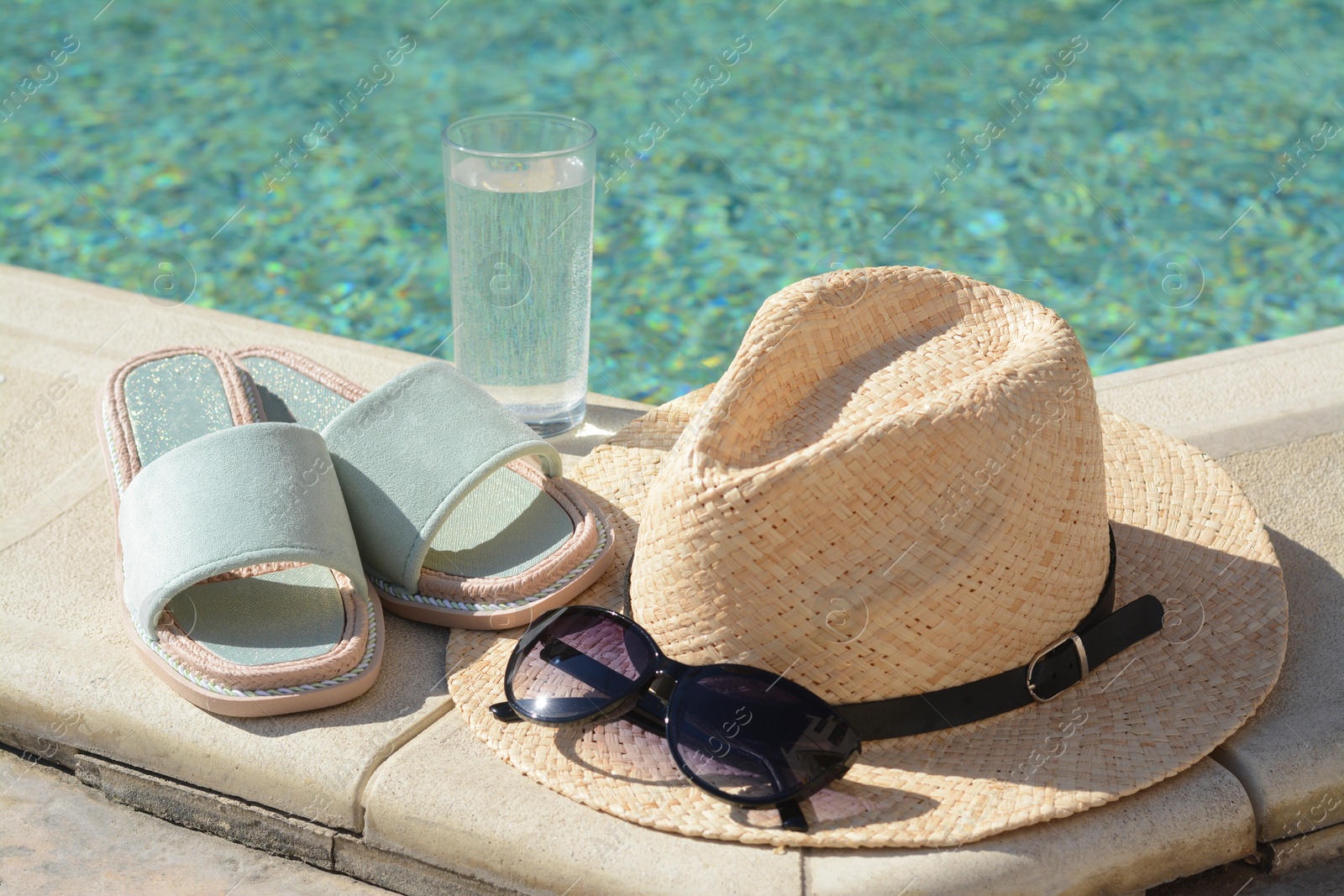 Photo of Stylish sunglasses, slippers, straw hat and glass of water at poolside on sunny day. Beach accessories