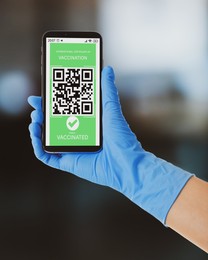 Image of Person in gloves holding smartphone with international certificate of vaccination on blurred background, closeup